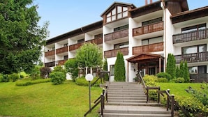 A multi-story hotel with a central staircase, surrounded by well-maintained greenery and a clear sky. - FEWO in the vacation park Oberallgäu with gr. Pool, bed linen, towels, final cleaning (Missen-Wilhams)