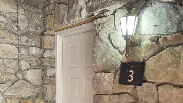 Una entrada amb parets de pedra, una porta blanca, un rètol amb el número 3 i un petit llum exterior.