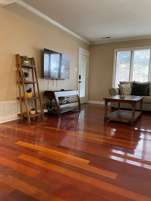 A living room with hardwood flooring, a TV mounted on the wall, a wooden ladder with plants, a sofa, and a coffee table.
