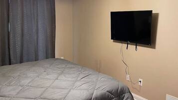 Een slaapkamer met een bed, een plafondventilator, een flatscreen televisie aan de muur en een raam met gordijnen.