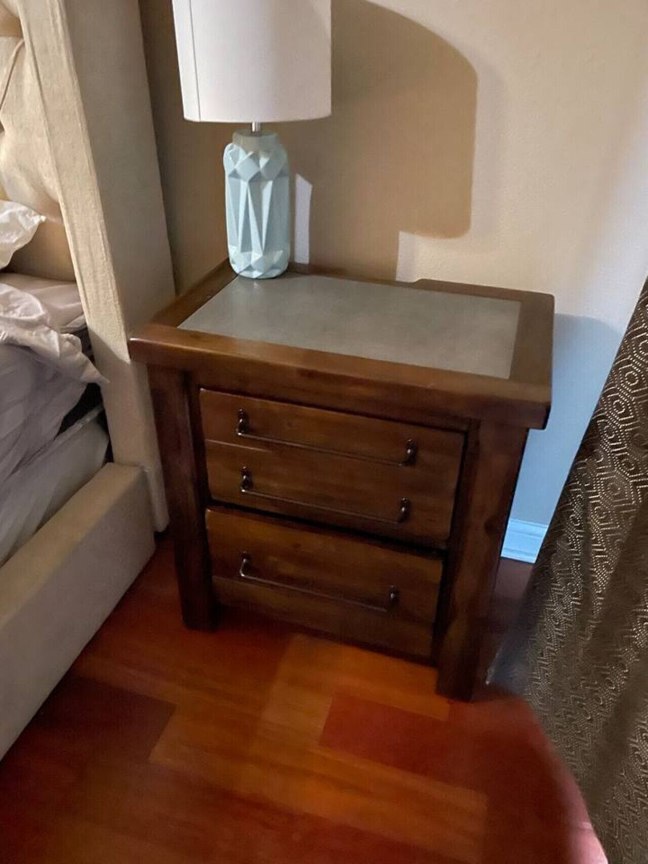 Ein hölzerner Nachttisch mit Lampe und Schublade neben einem Bett in einem Hotelzimmer.