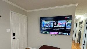A room with a wooden floor, a red bench, a white door, and a flat-screen TV mounted on the wall displaying news. - 3 Bed 1.5 Bath Pet Friendly Apt (Columbia)