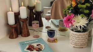 Ein Zimmer mit weißem Schreibtisch, Kerzen, einem Buch, einer Blumenvase und einem Fenster mit Blick.
