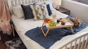 Ein Schlafzimmer mit einem weißen Bett, einem Nachttisch mit Lampe, einem Fenster mit Blick auf einen Zaun und einer Ablage mit Obst und Snacks auf dem Bett.