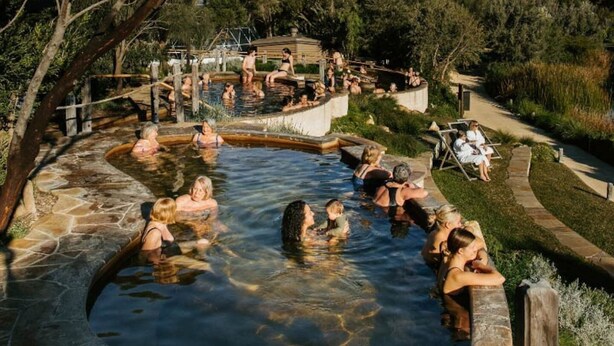 Ein Thermalgebiet mit Menschen, die im Wasser entspannen, umgeben von Steinbauten und Grünpflanzen.