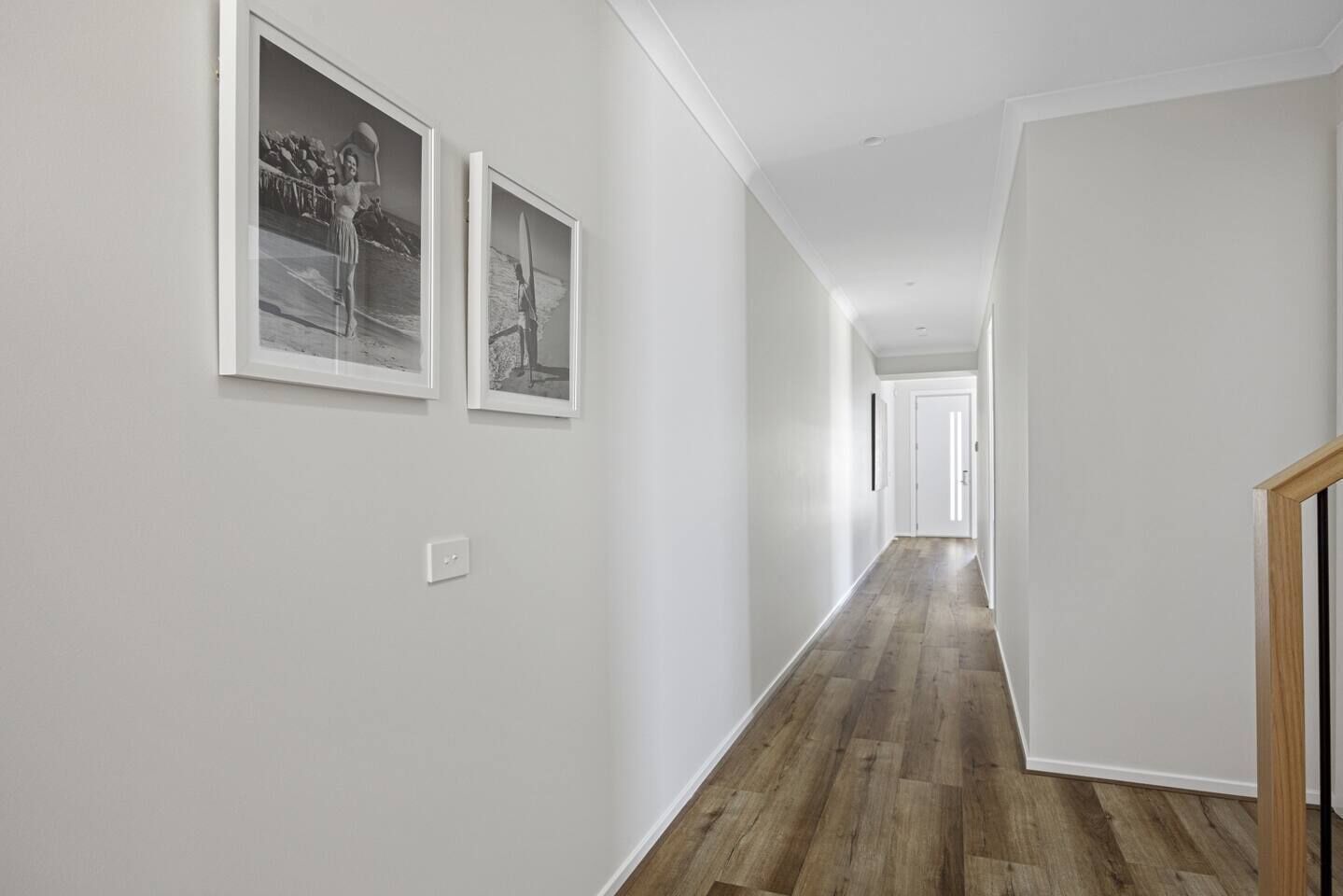 Un couloir avec un plancher en bois, des murs blancs et des tableaux encadrés sur le mur de gauche.