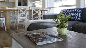 Una sala de estar moderna con una revista sobre una mesa de centro de madera, una planta verde y un cojín con la inscripción 'hogar dulce hogar' en un sofá gris.