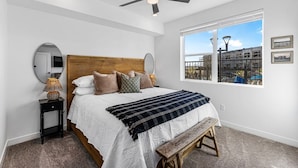Un dormitorio con una cama grande, cabecera de madera, ventilador de techo, mesita de noche con lámpara, ventana con vista a la ciudad y un banco.