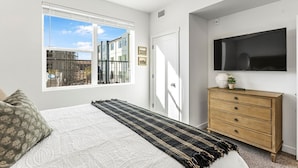 Un dormitorio con una cama grande, un aparador de madera, un televisor montado y una ventana con vista a la ciudad.