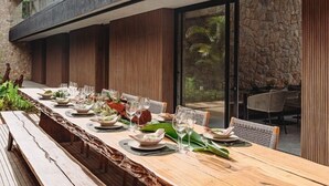 A long wooden dining table set with plates, glasses, and cutlery, flanked by stone walls and wooden paneling. - Pousada Maria do Mar (Fernando de Noronha)