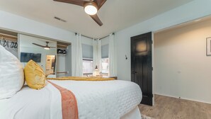 Une chambre à coucher avec un lit, un ventilateur de plafond, une fenêtre avec des rideaux, une porte et un miroir.
