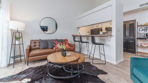 Un salon moderne avec un canapé en cuir marron, une table basse ronde en bois et un tapis en cuir noir. On aperçoit à l’arrière-plan un coin cuisine avec un bar et des chaises.