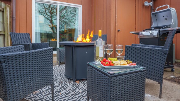 Une terrasse extérieure aménagée avec des meubles en osier, une table en verre sur laquelle sont disposés des plats, un barbecue et un foyer.