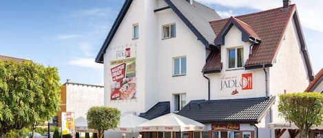 A two-story building with a restaurant named "Jadło" on the ground floor. There are outdoor seating areas with umbrellas, and a signboard advertising a 20% discount.