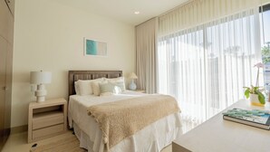 Una camera d'albergo con un letto grande, due comodini con lampade, una pianta sul comodino e un'ampia finestra con tende leggere.