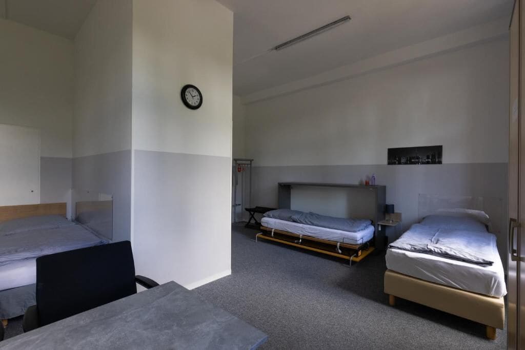 Ein Hotelzimmer mit zwei Einzelbetten, einem Schreibtisch, einem Stuhl, einer Wanduhr und einem kleinen Tisch mit einer Lampe.