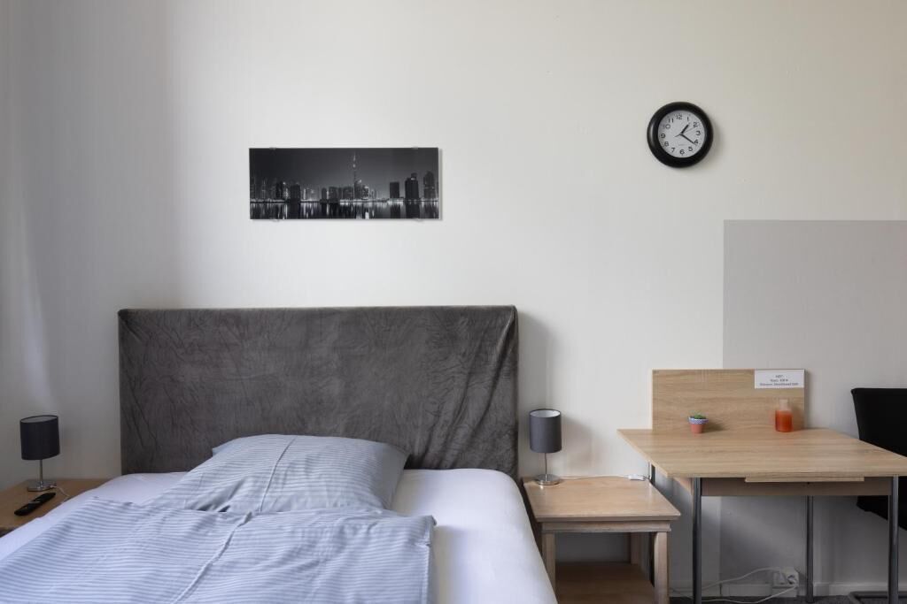 Ein Hotelzimmer mit Bett, Nachttisch mit Lampe, kleinem Tisch mit Pflanze und Flasche, einer Wanduhr und einem Stadtbild.