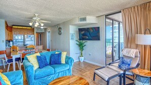 A living room with a blue sofa, a dining area with a table and chairs, a ceiling fan, and a large window with curtains. - Ocean Front, Private Balconies (Myrtle Beach)