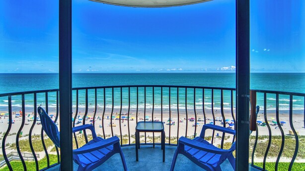 A balcony with two chairs and a table, overlooking a beach with umbrellas and people. - Ocean Front, Private Balconies (Myrtle Beach)
