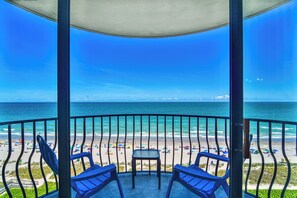 Un balcon avec deux chaises et une table, donnant sur une plage parsemée de parasols et fréquentée par des gens.