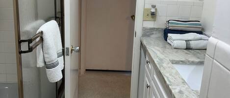 A narrow bathroom with a glass shower door, a white sink, and a countertop with towels.