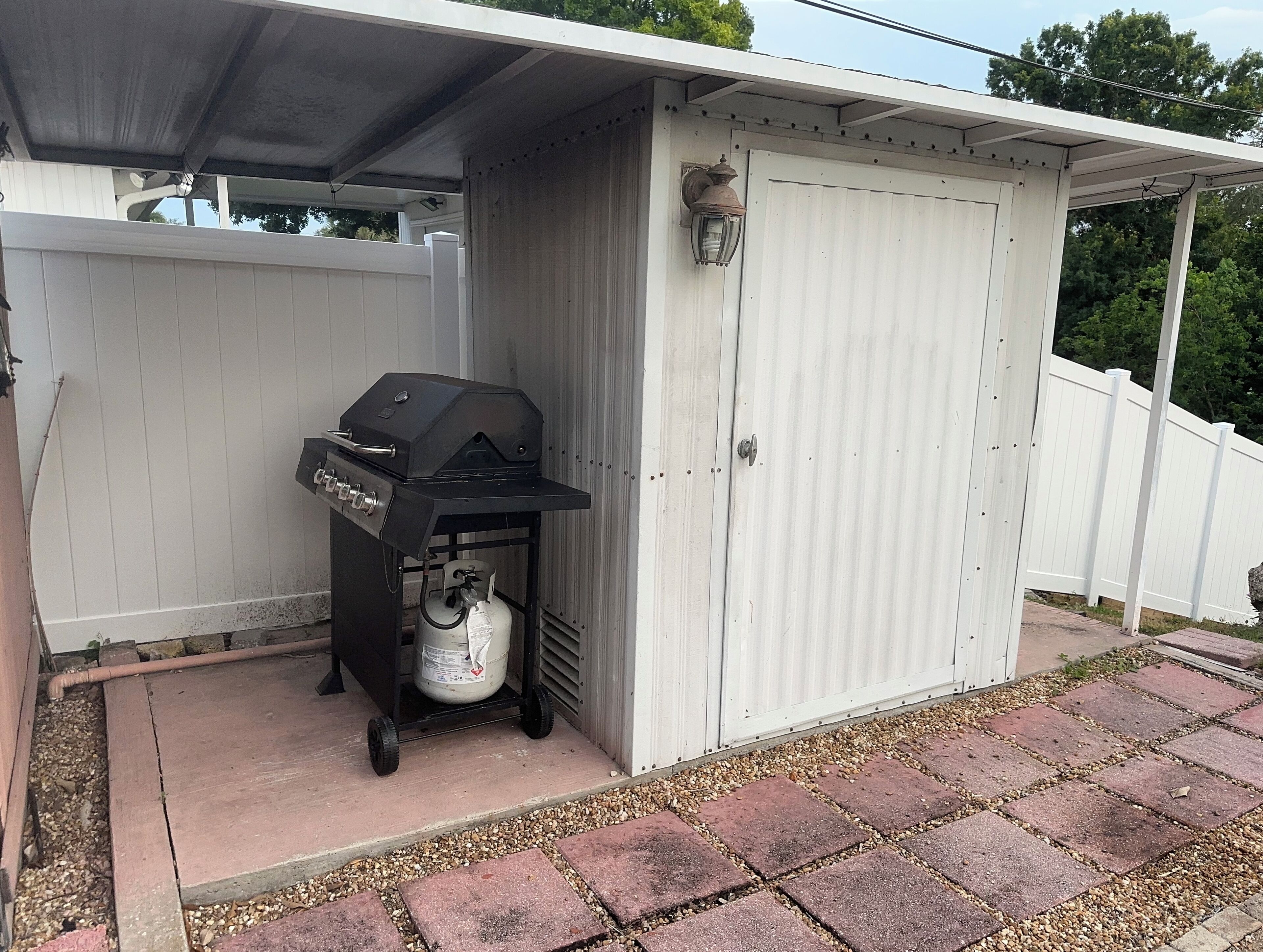 A barbecue grill with a propane tank under a covered area with a white fence.
