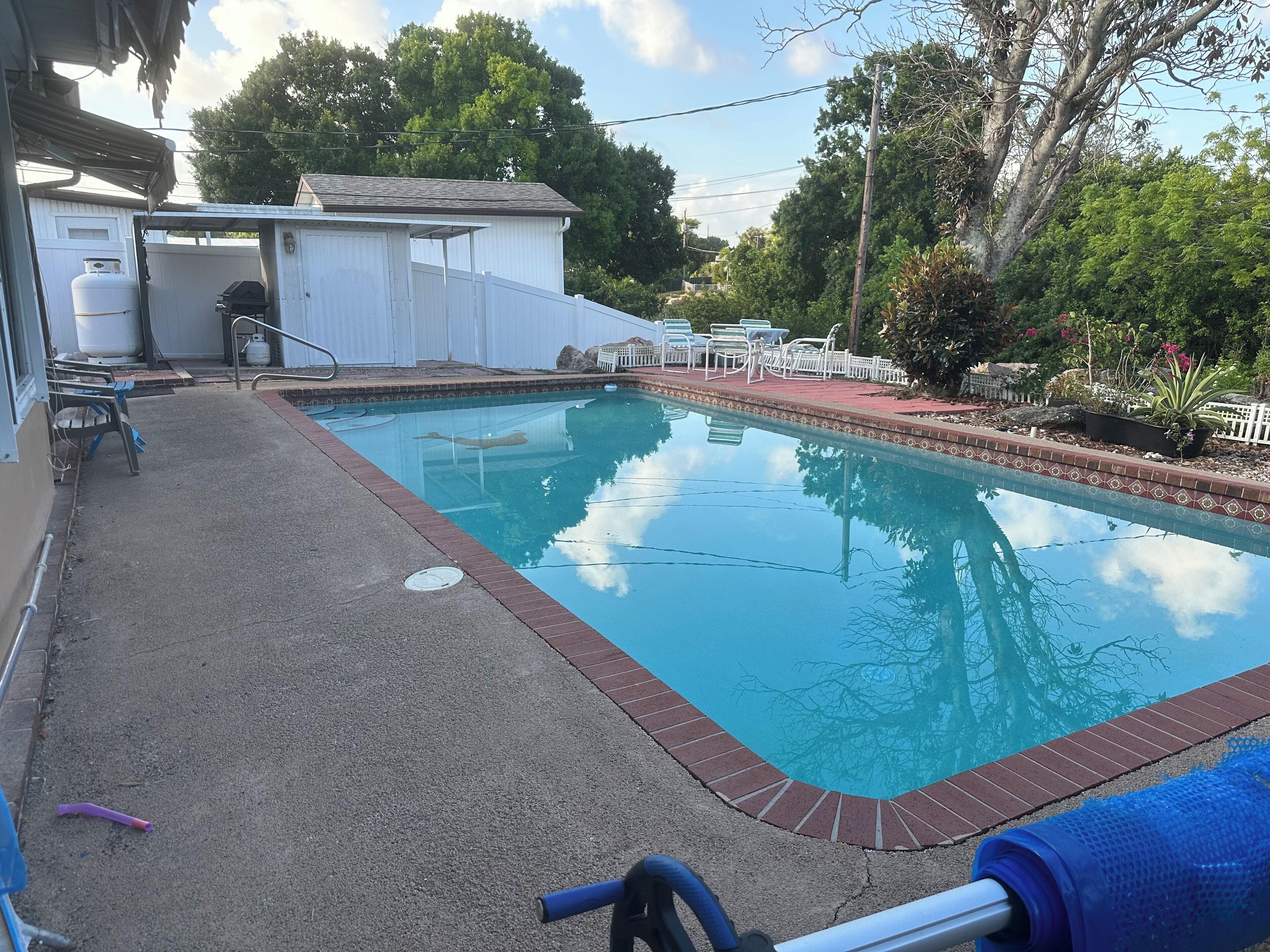 A swimming pool with a brick edge, surrounded by a patio with chairs and a barbecue grill.