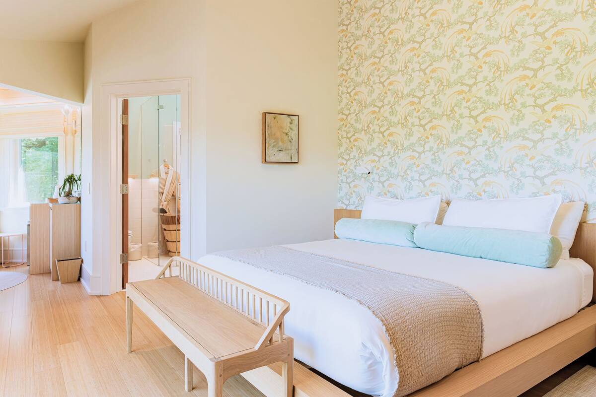 Un dormitorio con cama, un banco, un espejo y una puerta que da al baño.