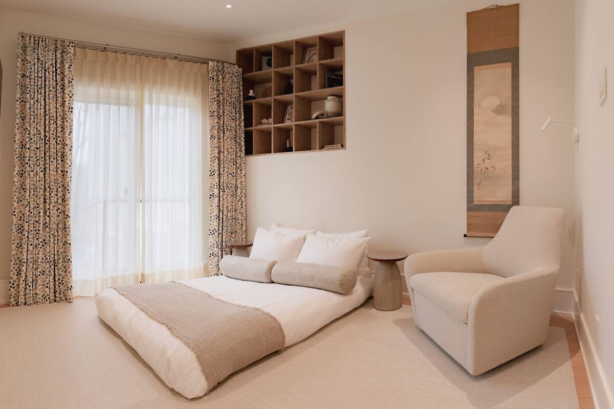 Un dormitorio con una cama, una silla, una estantería con objetos decorativos y un cuadro en la pared.