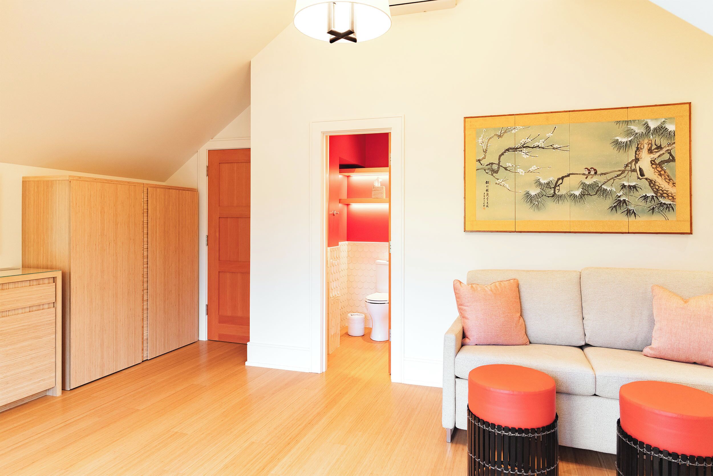 A room with a wooden wardrobe, a sofa with orange cushions, a painting on the wall, and a ceiling light fixture.