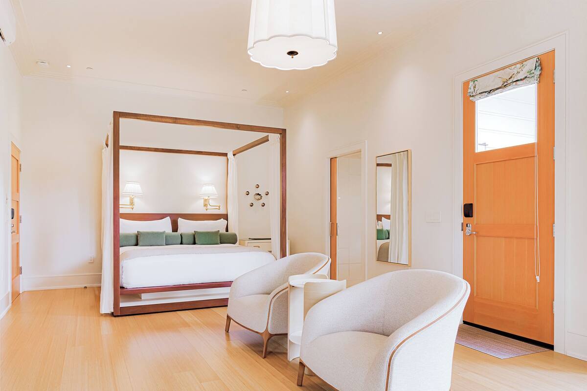 Un dormitorio con una cama con dosel, dos sillones, una puerta de madera y un espejo grande.