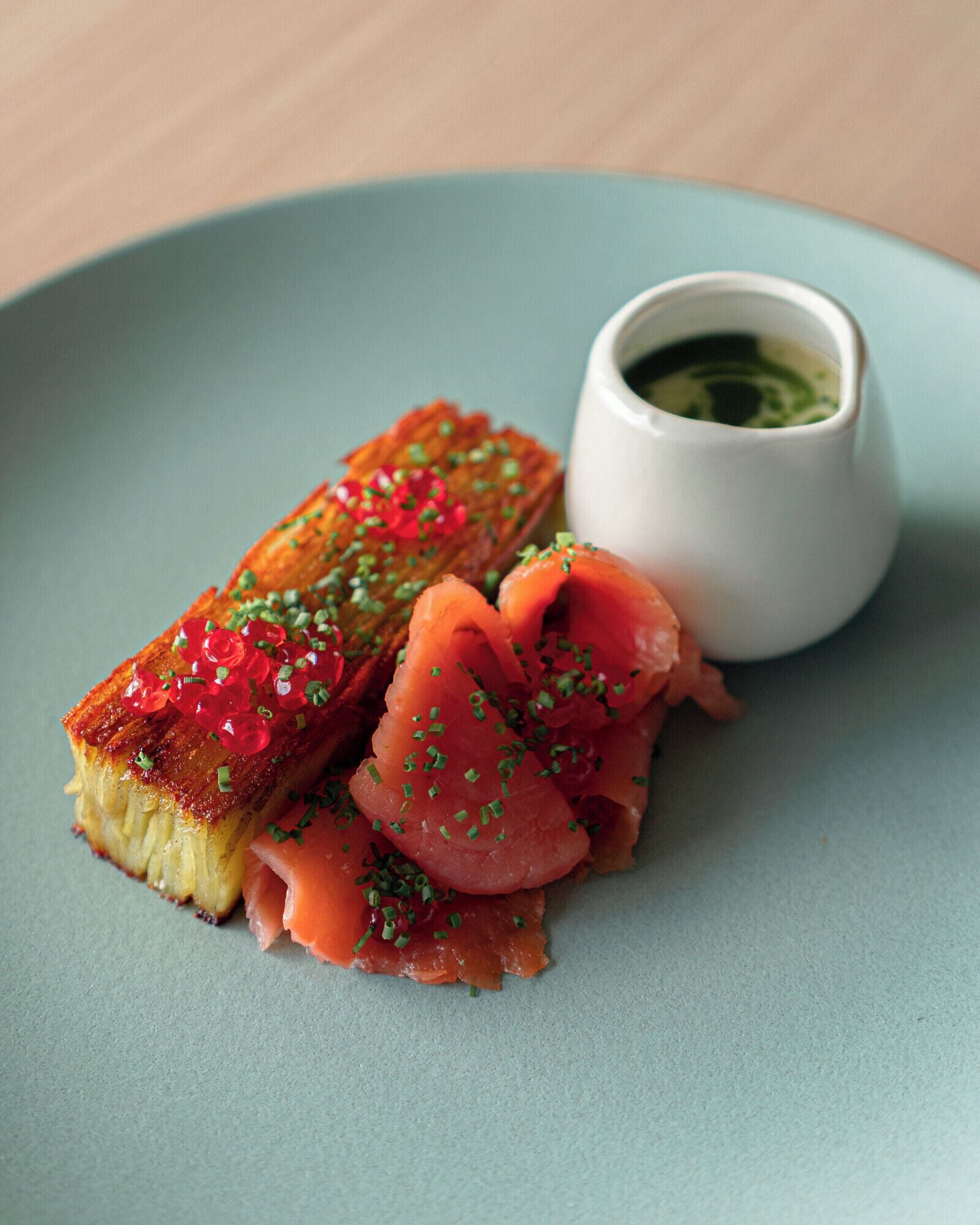 Un plato con un bocado de comida decorado con caviar rojo y hierbas verdes, junto a una pequeña taza blanca de salsa verde.