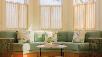 Un sofá verde con cojines estampados, una mesa redonda con un jarrón blanco y un ventanal con cortinas traslúcidas.