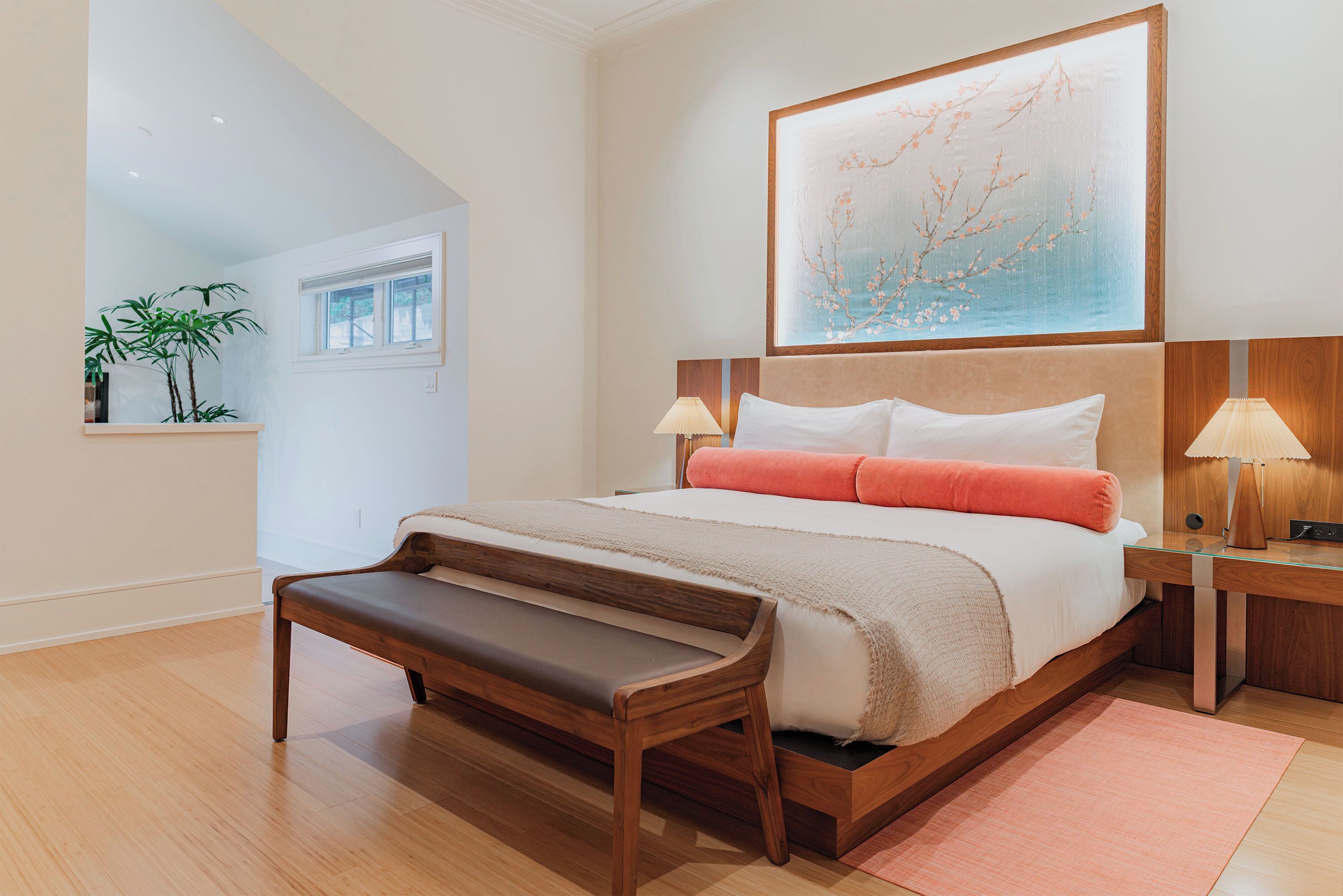 A modern bedroom with a large bed, bedside lamps, a bench, and a window with a view of greenery.