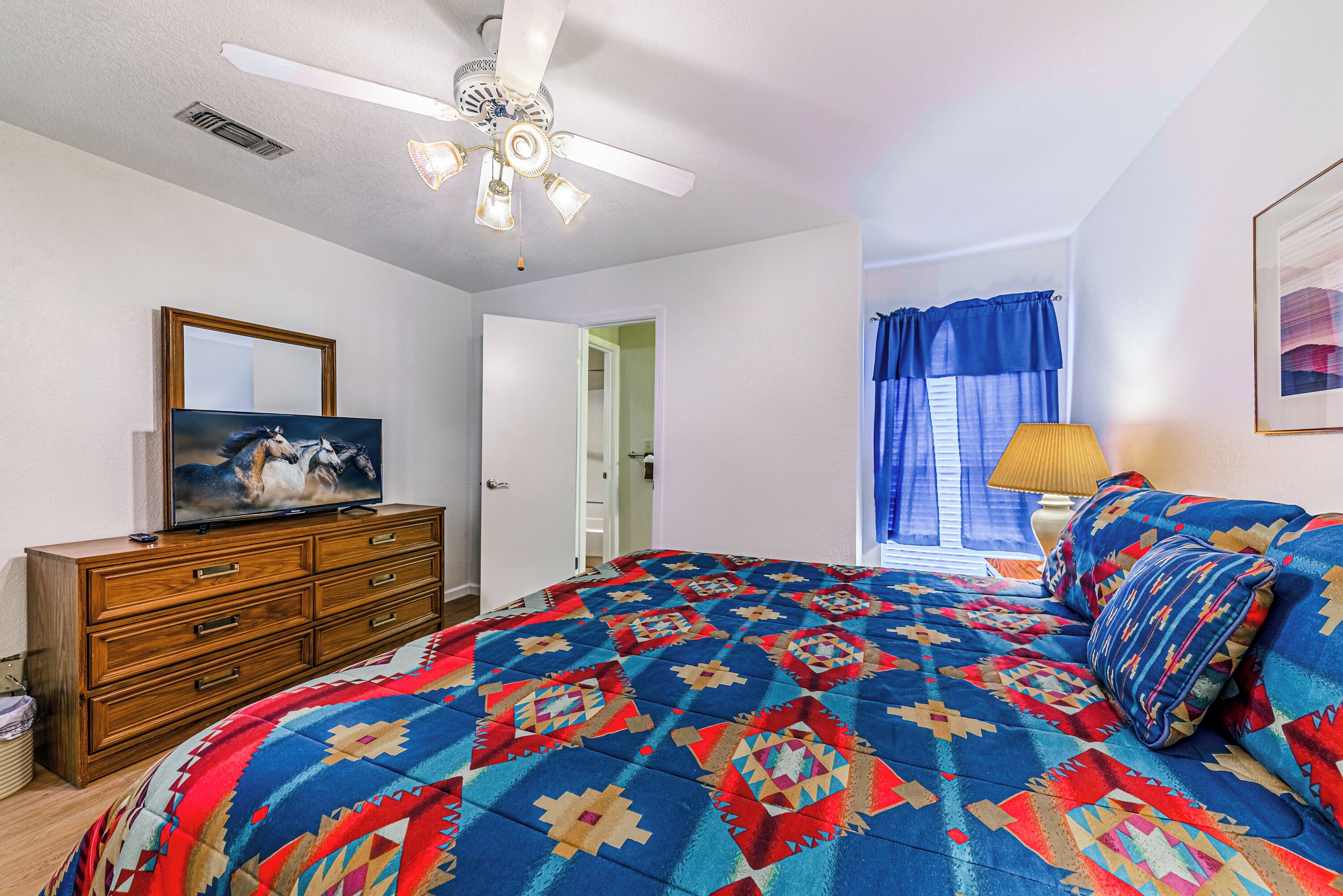 A bedroom with a bed, a dresser, a ceiling fan, a television, and a window with curtains.