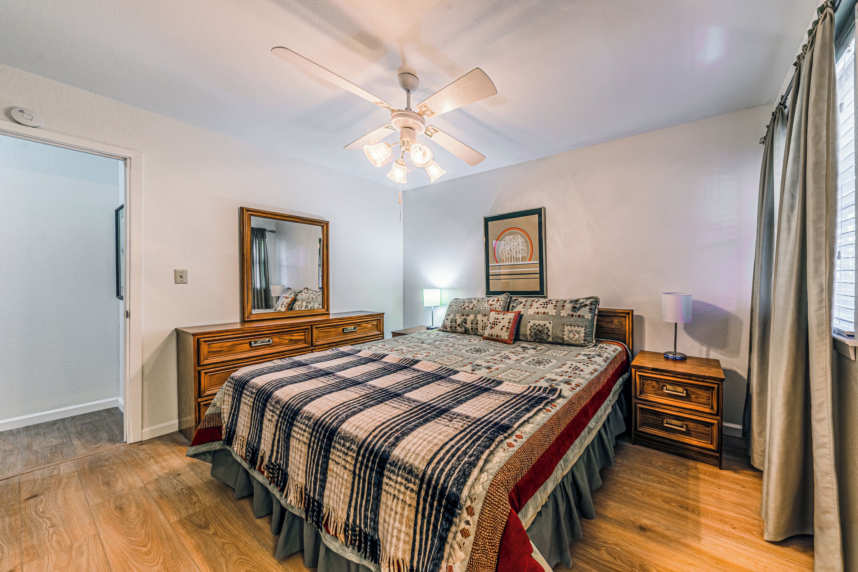 A bedroom with a bed, nightstands, a ceiling fan, a mirror, and a window with curtains.