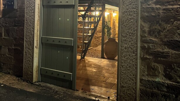 An arched stone entrance with a wooden door, a staircase inside, and a large vase. - Pietra Rossa (Chios)