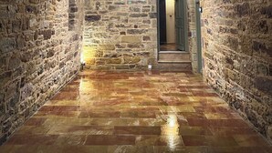 A stone-walled corridor with a tiled floor, a hanging light fixture, and a door at the end. - Pietra Rossa (Chios)