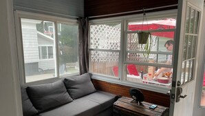 Una habitación con un sofá gris, un cofre de madera y una ventana con vista a un patio trasero con un paraguas rojo.