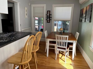 A dining area with a wooden table and chairs, a black puzzle on the table, and a window with a view of the outside.