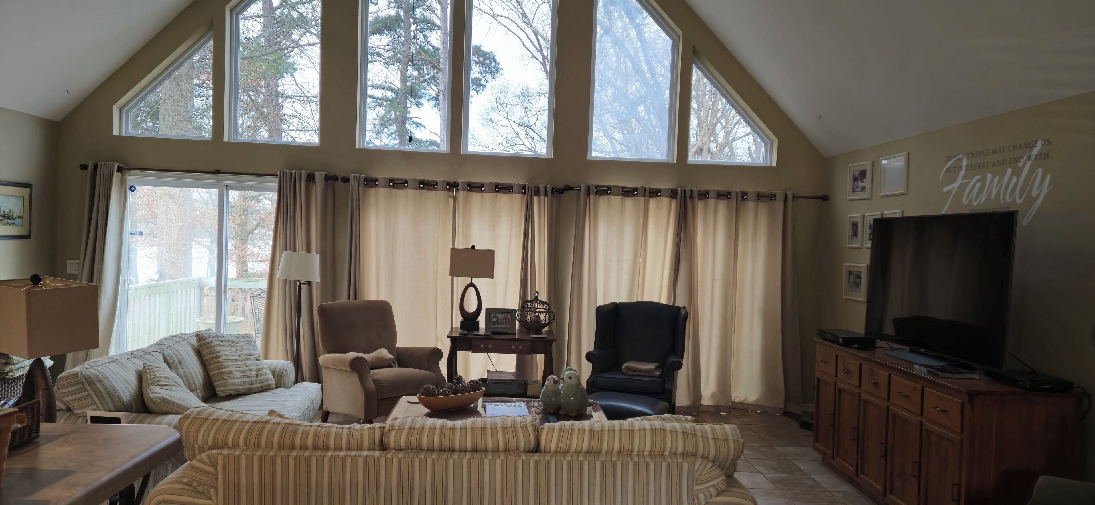 A spacious living room with high ceilings and large windows, featuring a sofa, two armchairs, a television, and a cabinet.
