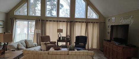 A spacious living room with high ceilings and large windows, featuring a sofa, two armchairs, a television, and a cabinet.
