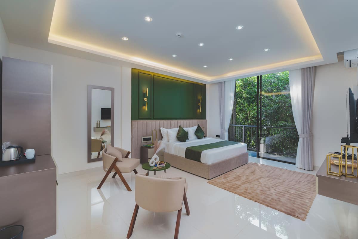 A modern hotel room with a large bed, a sitting area with a chair and table, a TV, and a balcony with a view of greenery.