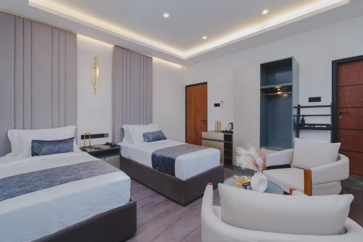 A modern hotel room with two beds, a seating area, and a small kitchenette.