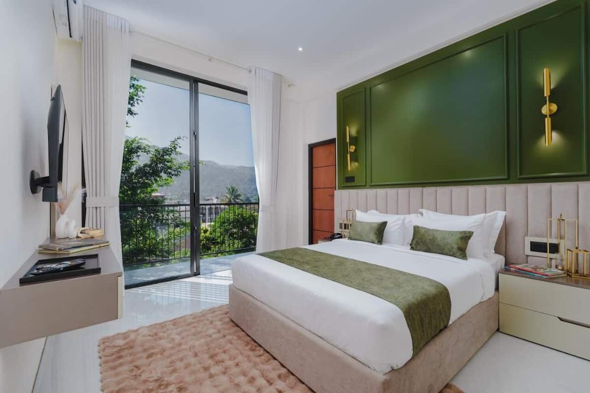 A modern bedroom with a large bed, a TV, and a balcony with a view of greenery and hills.