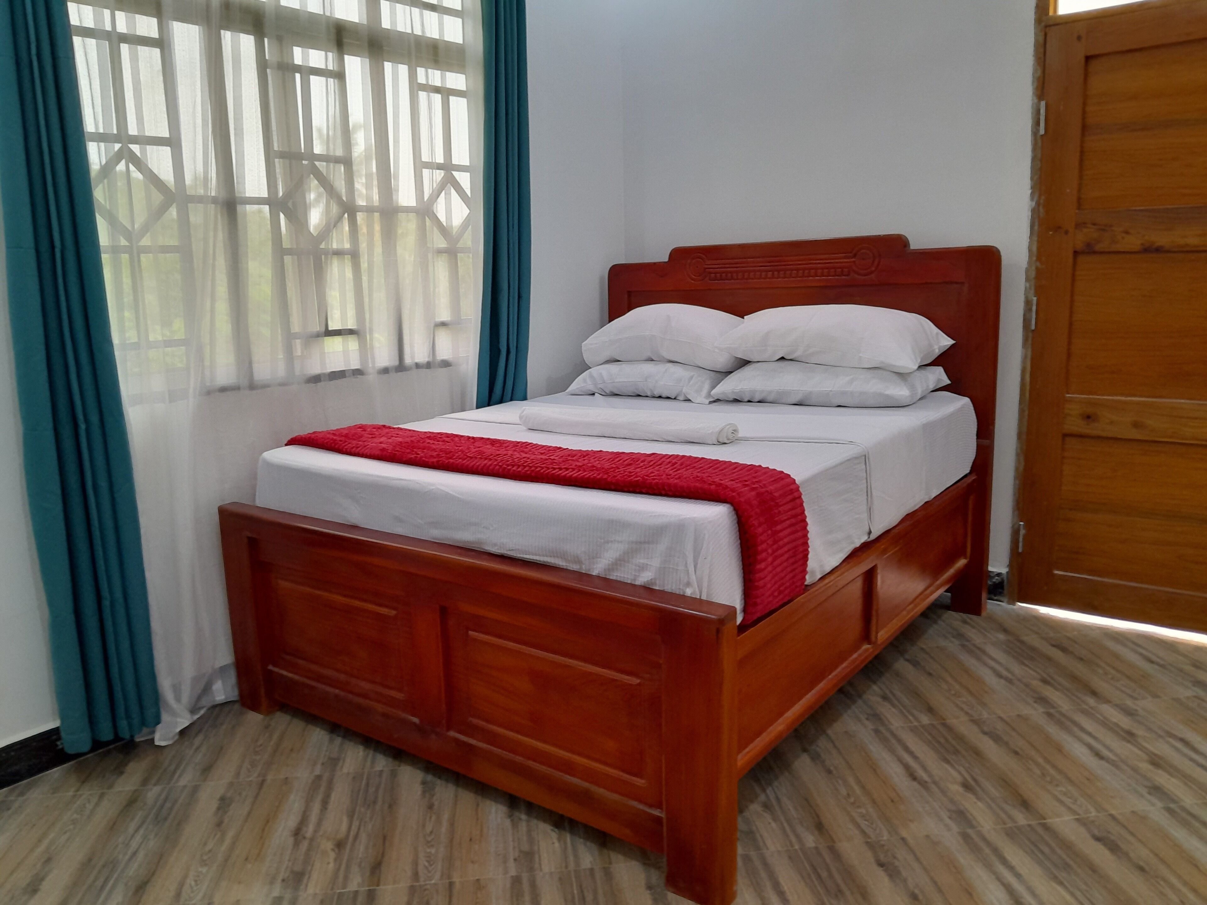 A wooden bed with white bedding and a red blanket in a room with wooden flooring and a window with blue curtains.