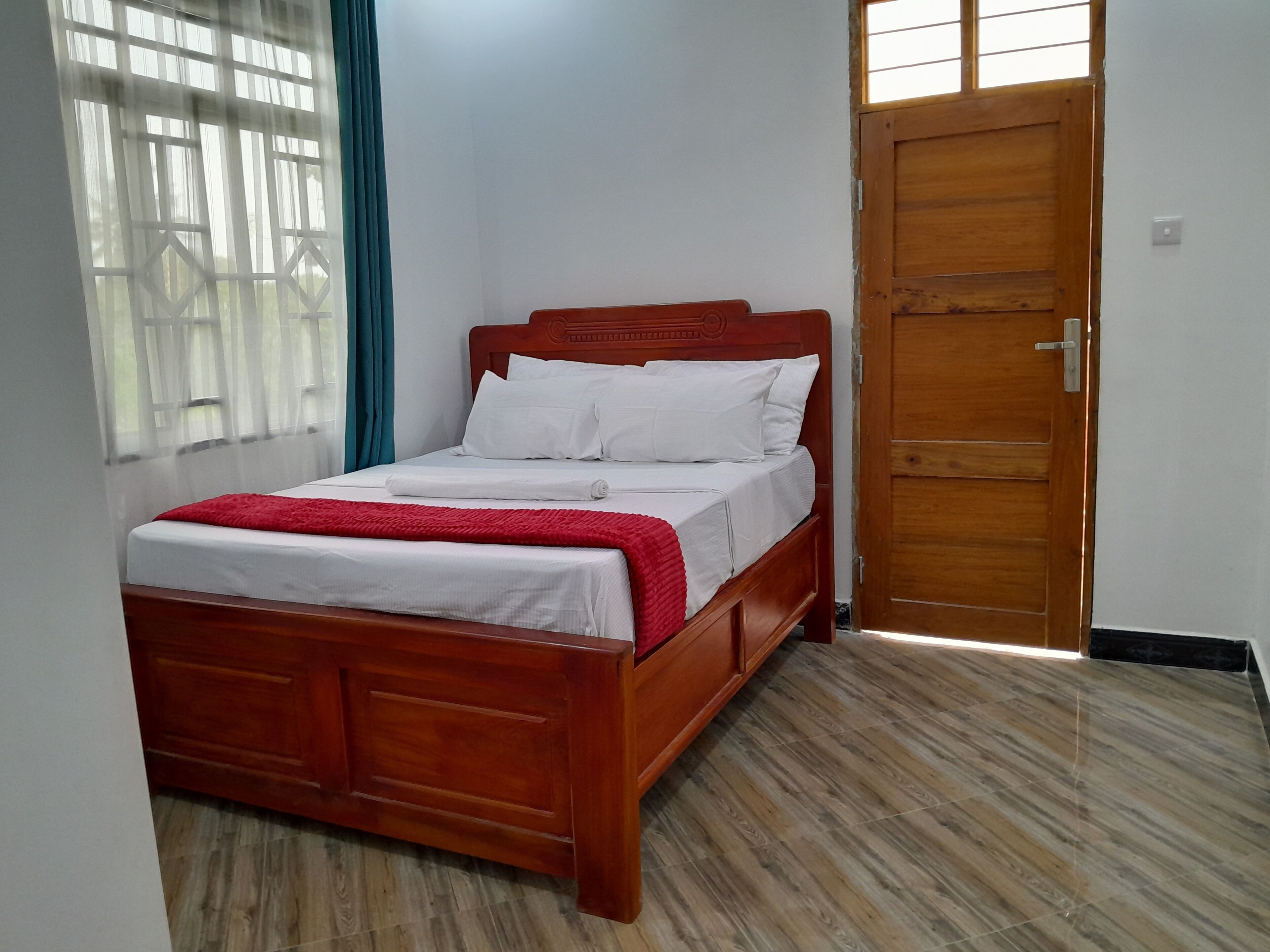 A neatly made bed with white linens and a red blanket, a wooden door, and a window with blue curtains.