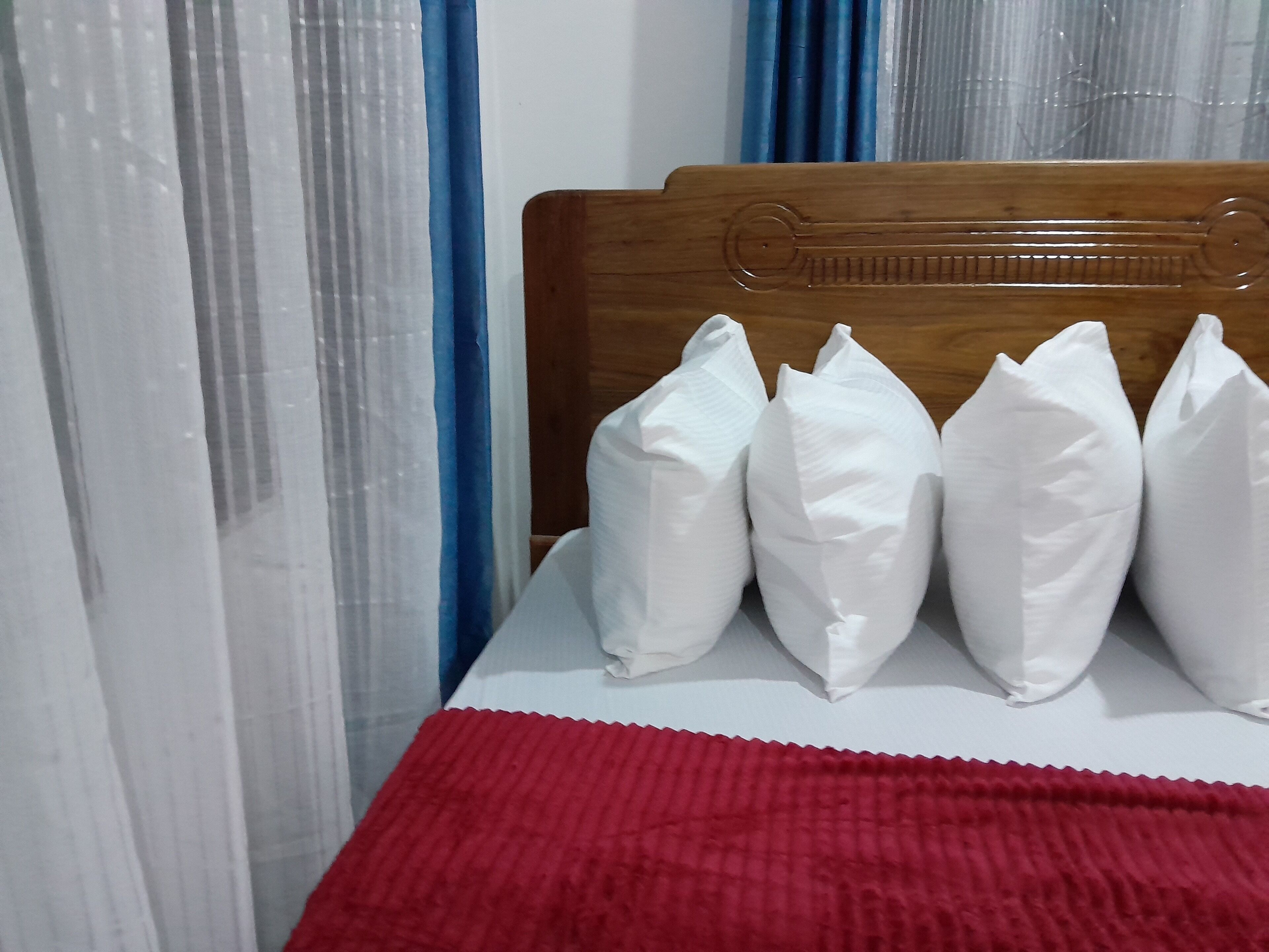 A bed with white pillows and a red blanket, a wooden headboard, and sheer curtains.