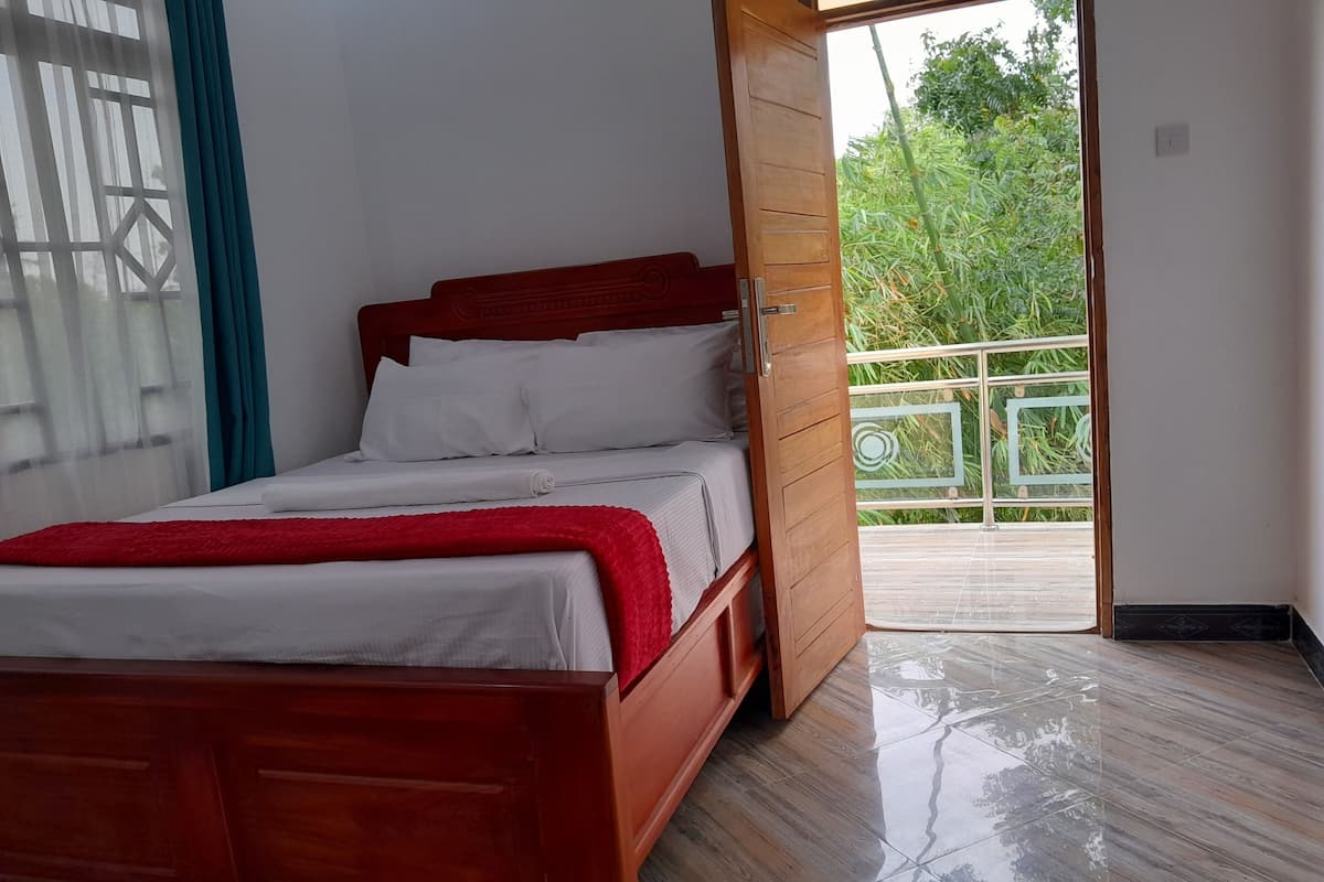 Una cama de madera con ropa de cama blanca y una manta roja en una habitación con suelo de madera y vistas a zonas verdes a través de una puerta abierta.