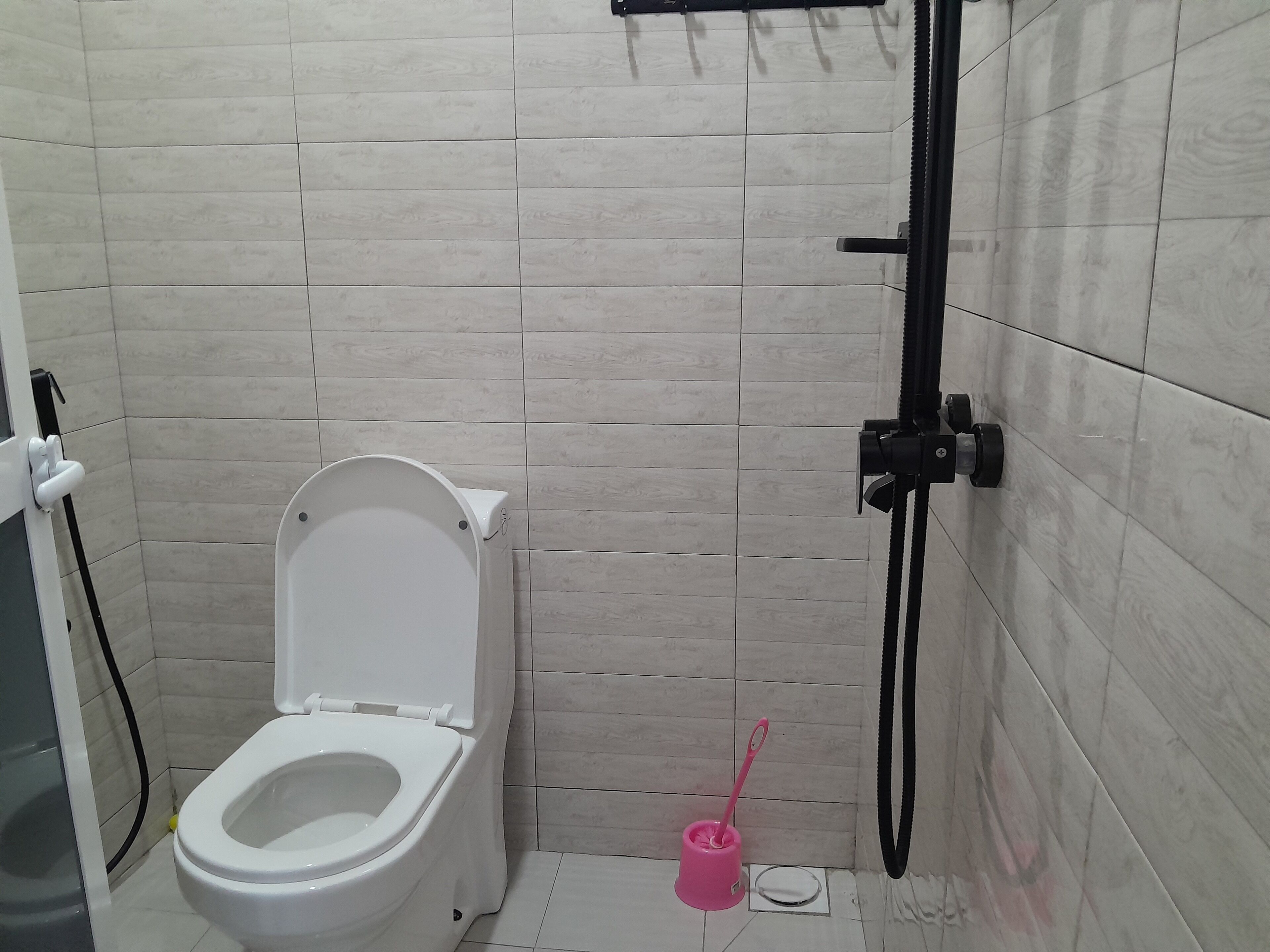 A small, modern bathroom with a white toilet, a shower head, and a pink cleaning brush.